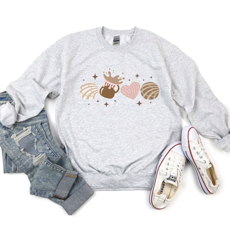 May include: A gray sweatshirt with a graphic design of a brown chocolate pot with a splash of chocolate, a pink heart-shaped pastry, and two brown shell-shaped pastries. The graphic is surrounded by small white stars.