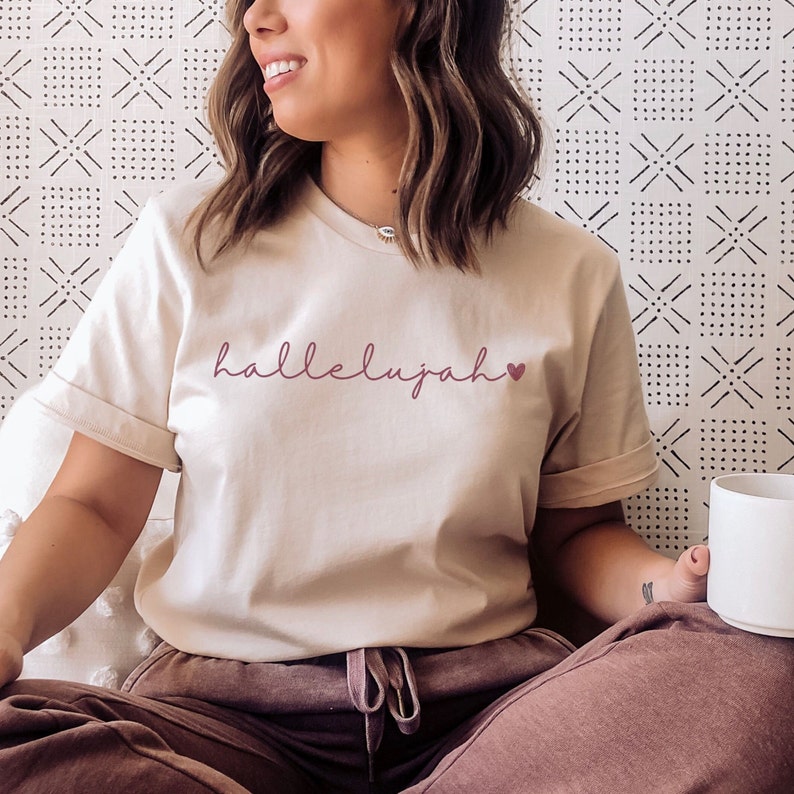 May include: A woman wearing a light beige t-shirt with the word "hallelujah" in pink script and a small pink heart.