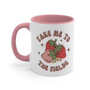 May include: White ceramic mug with a pink rim and handle. The mug features a graphic of two strawberries with green leaves and white flowers. The text "Take Me To The Fields" is printed in brown letters.