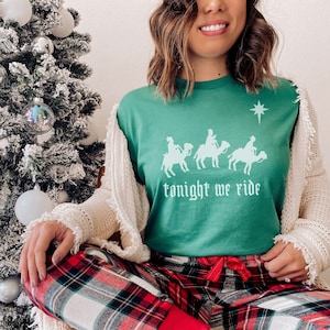 May include: A green t-shirt with a white graphic of the three wise men riding camels and the text "tonight we ride". The person wearing the shirt is also wearing red and black plaid pajama pants.
