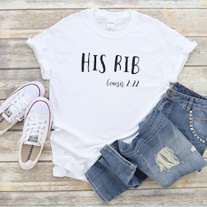 May include: White t-shirt with black text that reads "HIS RIB Genesis 2:22". The shirt is laid flat on a wooden surface with a pair of white sneakers and blue jeans.