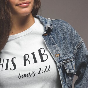 May include: White t-shirt with black text that reads "HIS RIB Genesis 2:22". The shirt is being worn by a person with long brown hair and a denim jacket.