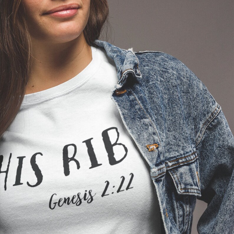 May include: White t-shirt with black text that reads "HIS RIB Genesis 2:22". The shirt is being worn by a person with long brown hair and a denim jacket.