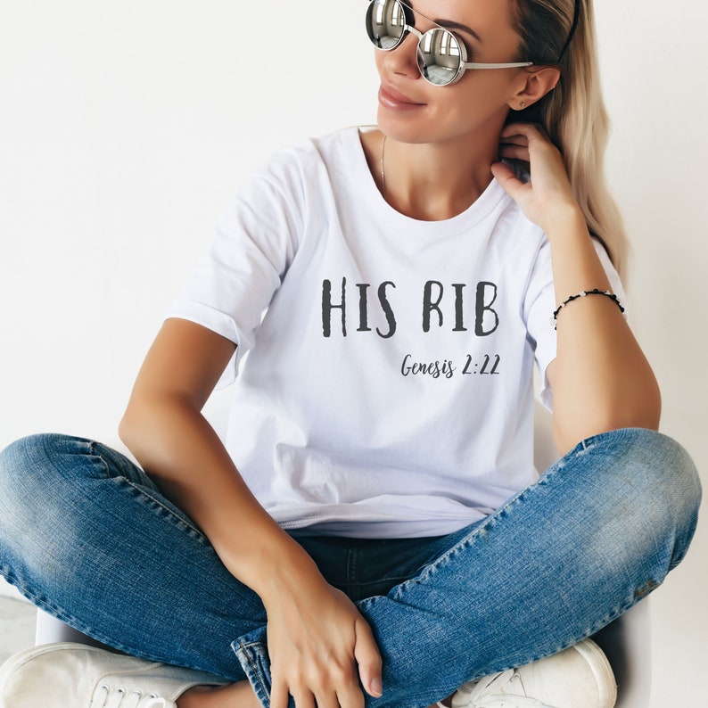 May include: A white t-shirt with the text "HIS RIB" and "Genesis 2:22" printed on it. The shirt is being worn by a person with blonde hair and sunglasses.