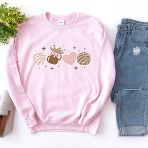 May include: A pink sweatshirt with a graphic of a brown mug with a chocolate drink spilling out, a brown and white concha, a pink and white heart-shaped concha, and a brown and white round concha. The graphic is surrounded by small brown stars.