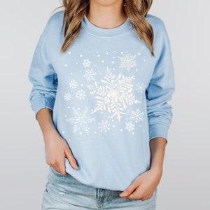 May include: A light blue sweatshirt with a white snowflake design. The snowflakes are arranged in a cluster and are surrounded by small white dots.