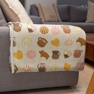 May include: A cream-colored fleece throw blanket with a repeating pattern of Mexican pastries, including conchas, pan dulce, and churros, along with brown clay pots and a brown pig.