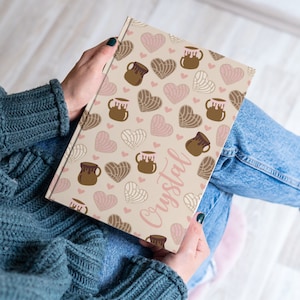 May include: A personalized notebook with a beige cover featuring a repeating pattern of brown and pink heart-shaped pastries and brown ceramic mugs. The name "Crystal" is written in pink cursive on the cover.