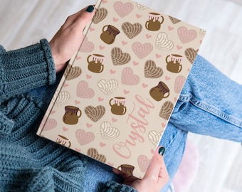 May include: A personalized notebook with a beige cover featuring a repeating pattern of brown and pink heart-shaped pastries and brown ceramic mugs. The name "Crystal" is written in pink cursive on the cover.