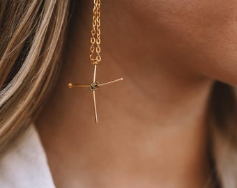 Może przedstawiać: Złote kolczyki z motywem krzyża. Kolczyki wiszą na delikatnym łańcuszku i mają unikalny, wiązany środek. Ramiona krzyża rozchodzą się na zewnątrz, tworząc nowoczesny i stylowy wygląd. Kolczyki są wyrazistym elementem biżuterii.
