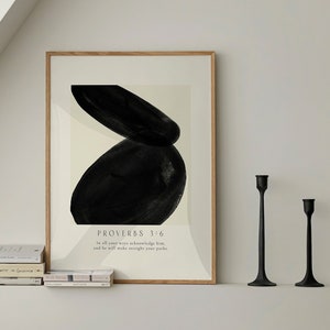 May include: A framed print with a black abstract design and the text "PROVERBS 3:6 To all your ways acknowledge him, and he will make straight your paths." The print is on a white background and is framed in a light wood frame. The print is sitting on a white shelf with a stack of books and two black candle holders.