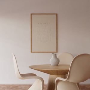 May include: A framed print of Psalm 23, a passage from the Bible, hangs on a light beige wall. The print is in a simple, modern style with a light brown frame. A wooden table with a white ceramic pitcher sits in front of the print. Three beige chairs surround the table.