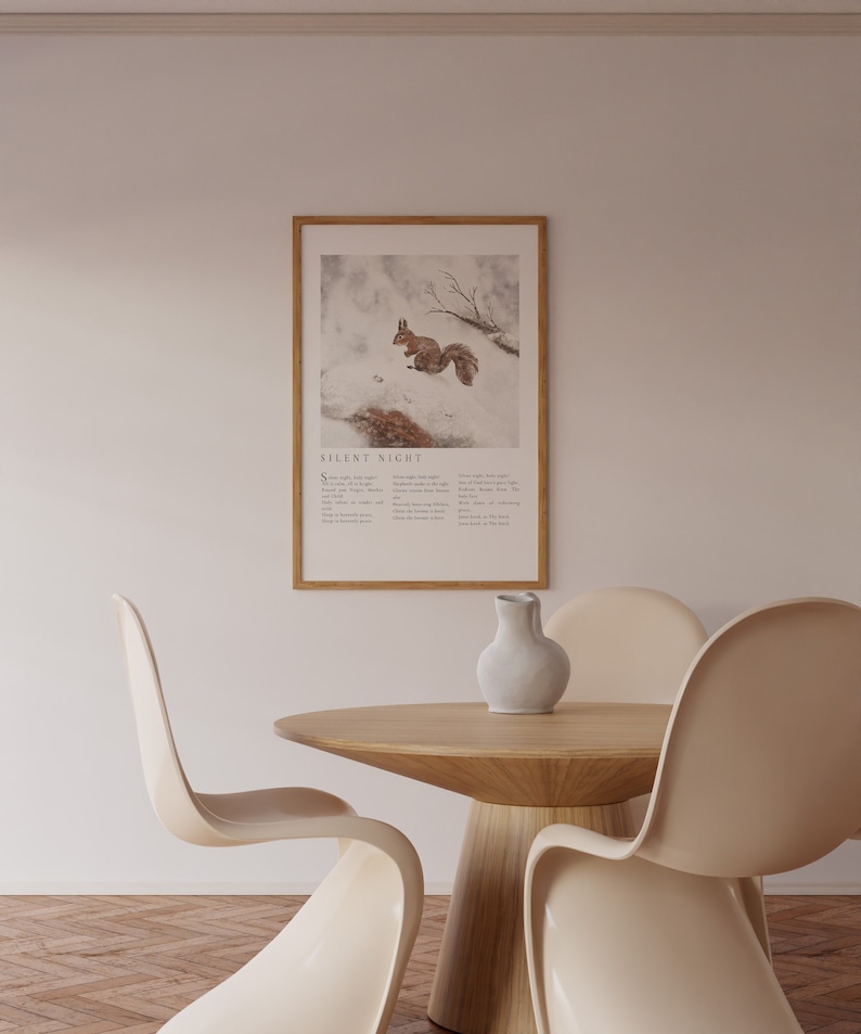 May include: A framed print with a winter scene featuring a squirrel in the snow. The print includes the lyrics to the Christmas carol "Silent Night". The print is hanging on a white wall above a wooden table with three beige chairs.