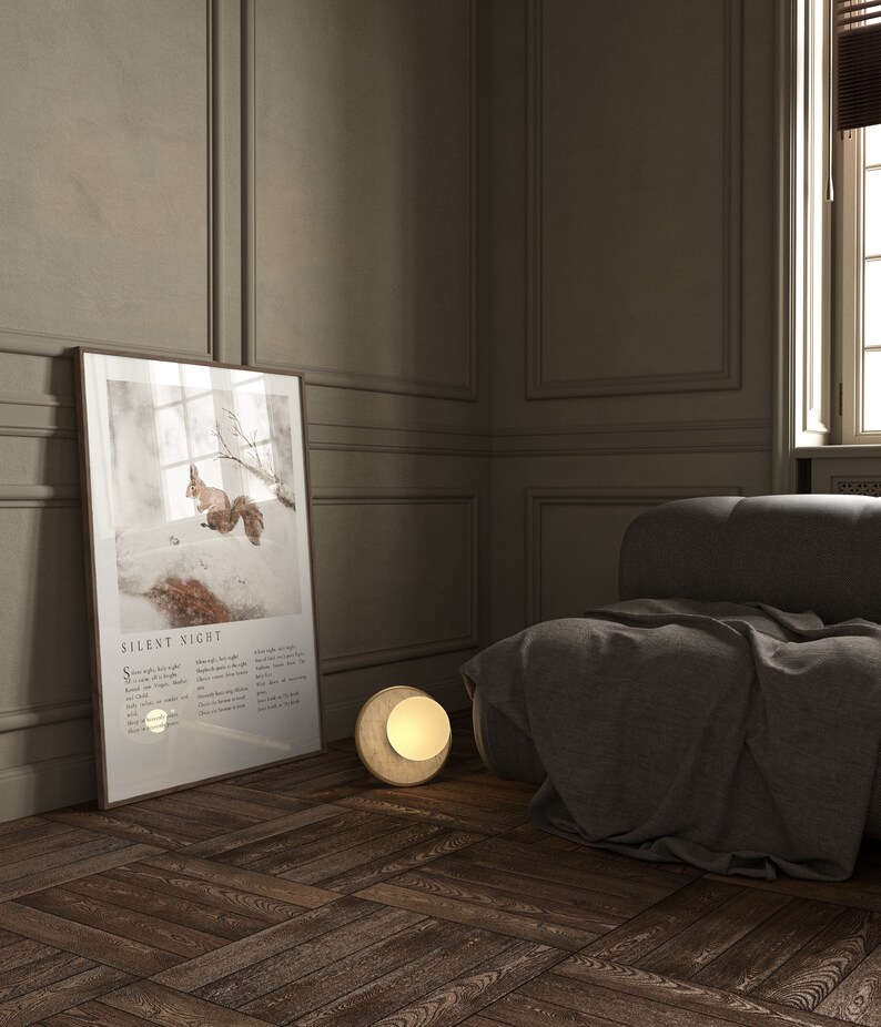 May include: A framed print of a squirrel in a snowy forest with the text "Silent Night" and lyrics below the image. A round, gold-colored lamp sits on the wooden floor next to a gray couch.