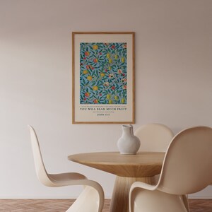 May include: A framed print with a blue background and a floral pattern featuring yellow lemons, red apples, and green leaves. The print includes the text "You will bear much fruit" and "John 15:5". The print is hanging on a white wall above a wooden table with two white chairs.