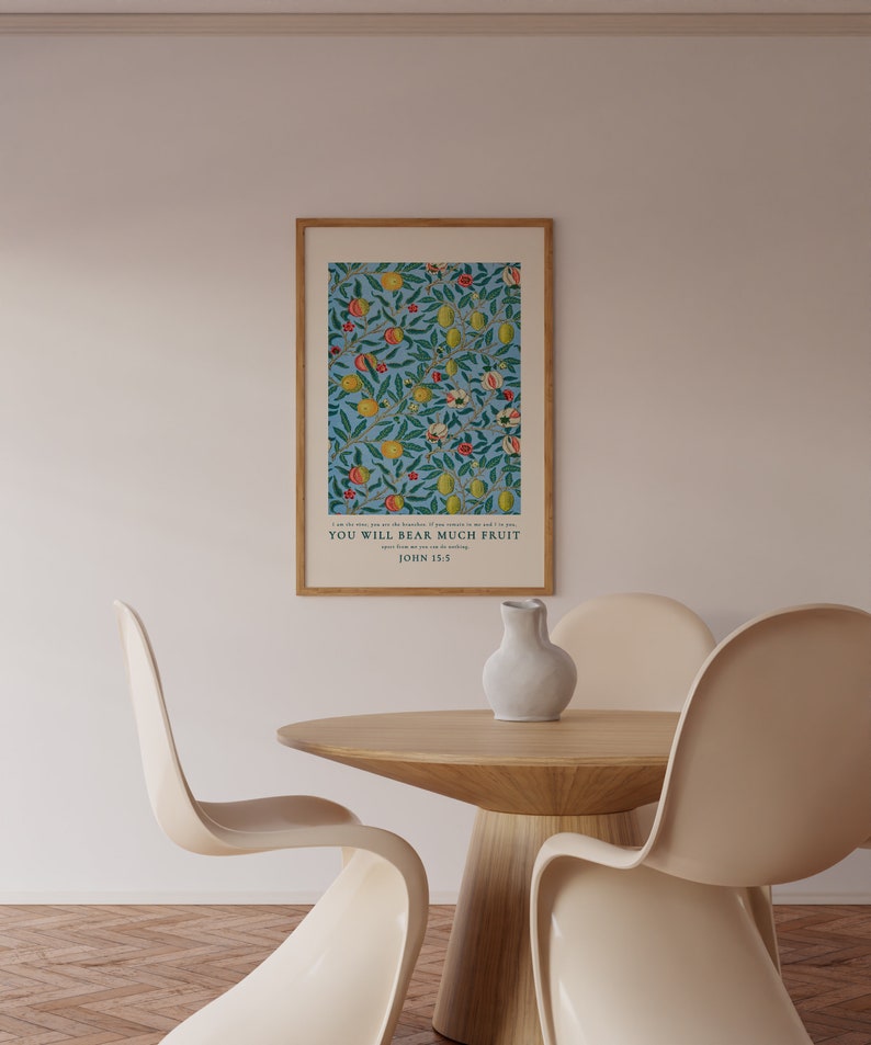 May include: A framed print with a blue background and a floral pattern featuring yellow lemons, red apples, and green leaves. The print includes the text "You will bear much fruit" and "John 15:5". The print is hanging on a white wall above a wooden table with two white chairs.