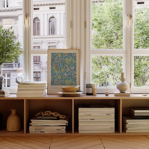 May include: A wooden shelf unit with three shelves, each filled with stacks of magazines. A framed print with a floral pattern and the text "You Will Be" is on the top shelf. A wooden bowl, a glass jar, and a ceramic vase are also on the top shelf.