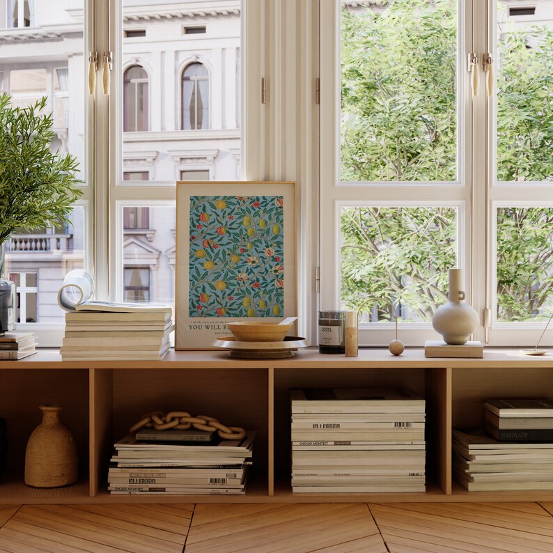 May include: A wooden shelf unit with three shelves, each filled with stacks of magazines. A framed print with a floral pattern and the text "You Will Be" is on the top shelf. A wooden bowl, a glass jar, and a ceramic vase are also on the top shelf.