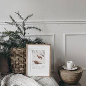 May include: A framed print of a squirrel in the snow with the text "Silent Night" below the image. The print is on a white shelf with a white cup and saucer on a wooden table next to it. A green plant in a wicker basket is on the left side of the image.