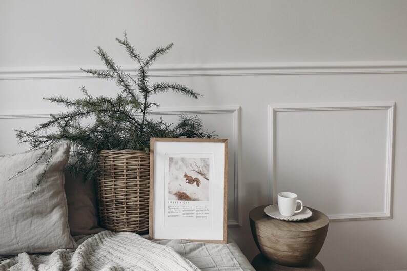 May include: A framed print of a squirrel in the snow with the text "Silent Night" below the image. The print is on a white shelf with a white cup and saucer on a wooden table next to it. A green plant in a wicker basket is on the left side of the image.
