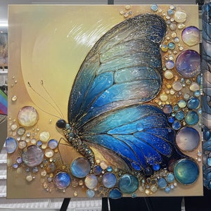 Puede incluir: Pintura sobre lienzo cuadrada de una mariposa con alas azules y doradas. La mariposa está rodeada de formas iridiscentes en forma de burbujas. El fondo es un degradado de amarillo y oro. La obra de arte se exhibe en un caballete negro.