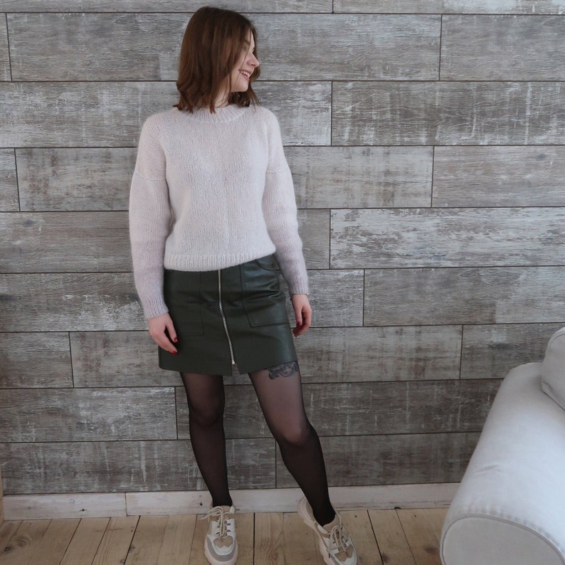 May include: A woman wearing a light pink fuzzy sweater and a dark green leather skirt. She is wearing black tights and white sneakers.