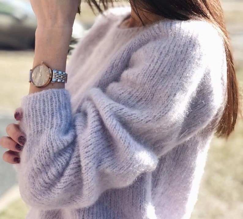 May include: A soft, light purple sweater with a loose fit and long sleeves. The sweater is made of a fuzzy, mohair-like yarn.