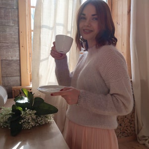 May include: A woman in a light pink sweater and a peach-colored pleated skirt holds a white cup and saucer. She is standing in front of a window with white curtains and a wooden wall behind her.