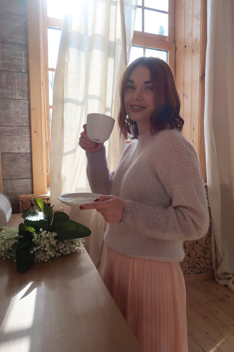 May include: A woman in a light pink sweater and a peach-colored pleated skirt holds a white cup and saucer. She is standing in front of a window with white curtains and a wooden wall behind her.
