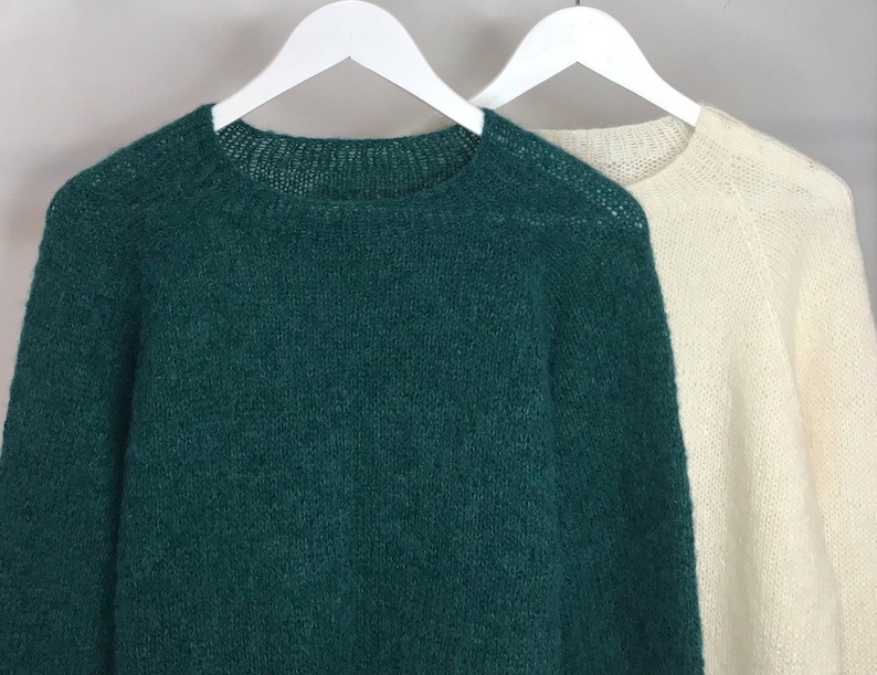 May include: Two sweaters, one green and one white, hanging on white hangers against a light gray background.