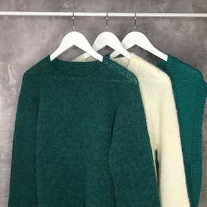 May include: Three sweaters hanging on white hangers. The sweater on the left is a dark green, the sweater in the middle is white, and the sweater on the right is a dark green.