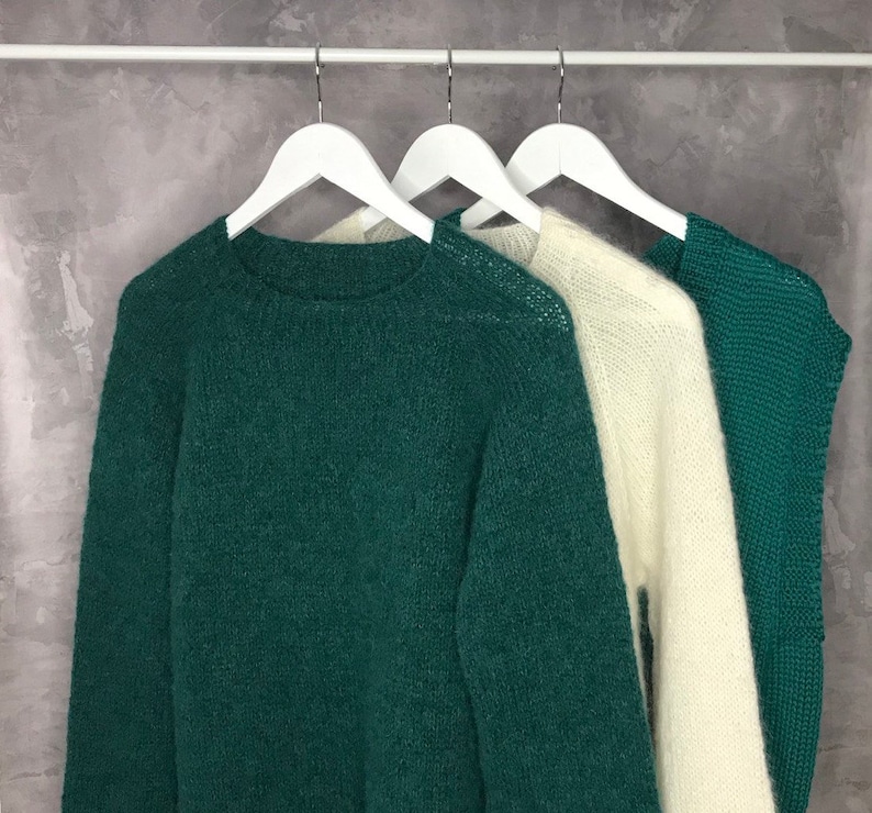 May include: Three sweaters hanging on white hangers. The sweater on the left is a dark green, the sweater in the middle is white, and the sweater on the right is a dark green.