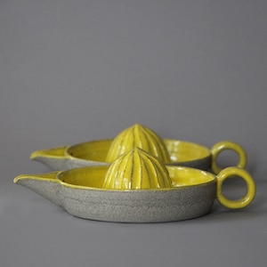 Könnte beinhalten: Zwei Zitruspressen aus Keramik. Die Entsafter haben eine graue Außenseite und eine leuchtend gelbe Innenseite. Jeder Entsafter hat einen Ausguss und einen Griff. Die Reibahlen sind gelb und gerippt.