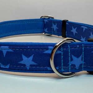 May include: A blue dog collar with a star pattern. The collar features a dark blue fabric with light blue stars, a silver metal D-ring, and a black plastic buckle. The collar is approximately 2.5 cm wide.
