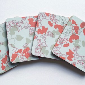 May include: Set of four square coasters with a floral design. The coasters feature a light blue background with a white outline of a floral pattern and pink flower accents.