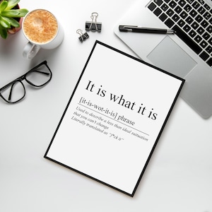 May include: A black framed print with the phrase "It is what it is" in bold, black text. The definition of the phrase is also included. The print is on a white surface with a laptop, coffee cup, glasses, and office supplies.