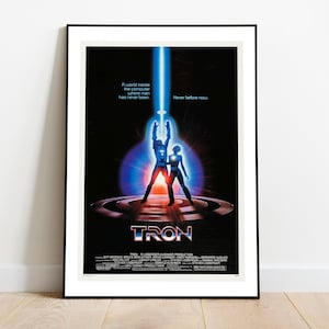 以下が含まれることがあります： 映画「トロン」のフレーム入りポスター。ポスターには、青い光線に照らされた未来的なスーツを着た2人の人物が描かれています。「トロン」のタイトルが下部に大きく表示され、追加のテキストとクレジットも表示されています。