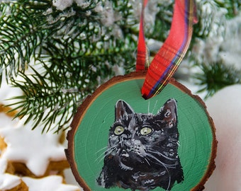 May include: A round wooden slice ornament featuring a hand-painted black cat on a solid green background. The ornament has a natural bark edge and is suspended by a red, green, and yellow plaid ribbon attached through a small metal eyelet.