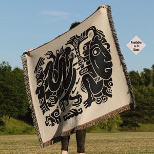 Pu&ograve; includere: Una coperta in tessuto beige con un design grafico in bianco e nero di figure stilizzate. Il design presenta forme e motivi geometrici. La coperta ha frange sui bordi.