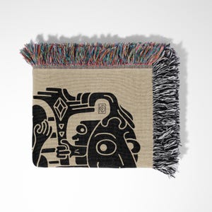 Pu&ograve; includere: Una coperta tessuta con un design grafico in bianco e nero di un viso stilizzato. La coperta ha un bordo a frangia con fili multicolori.