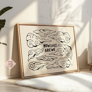 May include: A framed art print with a cream background and black cloud illustrations. The text "HOW LUCKY ARE WE" is displayed. The frame is a light wood colour. The artwork is a decorative piece.