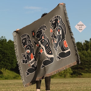 May include: A grey woven throw blanket with a black and orange design of three stylised figures. The figures are in a line and appear to be walking. The text "Available in 3 Sizes" is in the top right corner of the blanket.