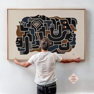 May include: Abstract art print in a wooden frame. The artwork features a geometric design with black, brown, and blue shapes on a cream background. A diamond shape is in the center. The art is held by a person.
