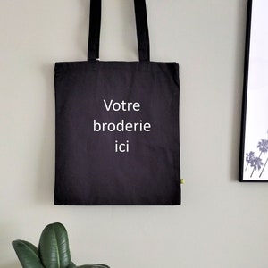 May include: Black tote bag with white text that reads "Votre broderie ici" hanging on a wooden hanger against a light green wall.