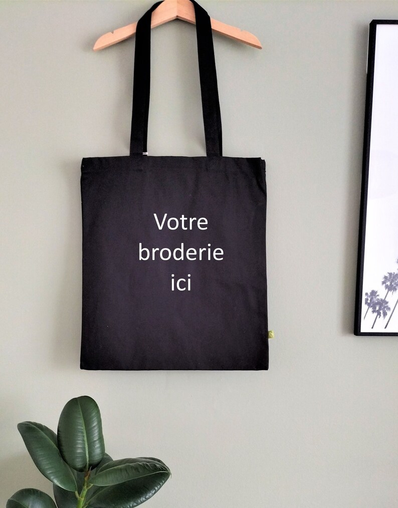 May include: Black tote bag with white text that reads "Votre broderie ici" hanging on a wooden hanger against a light green wall.