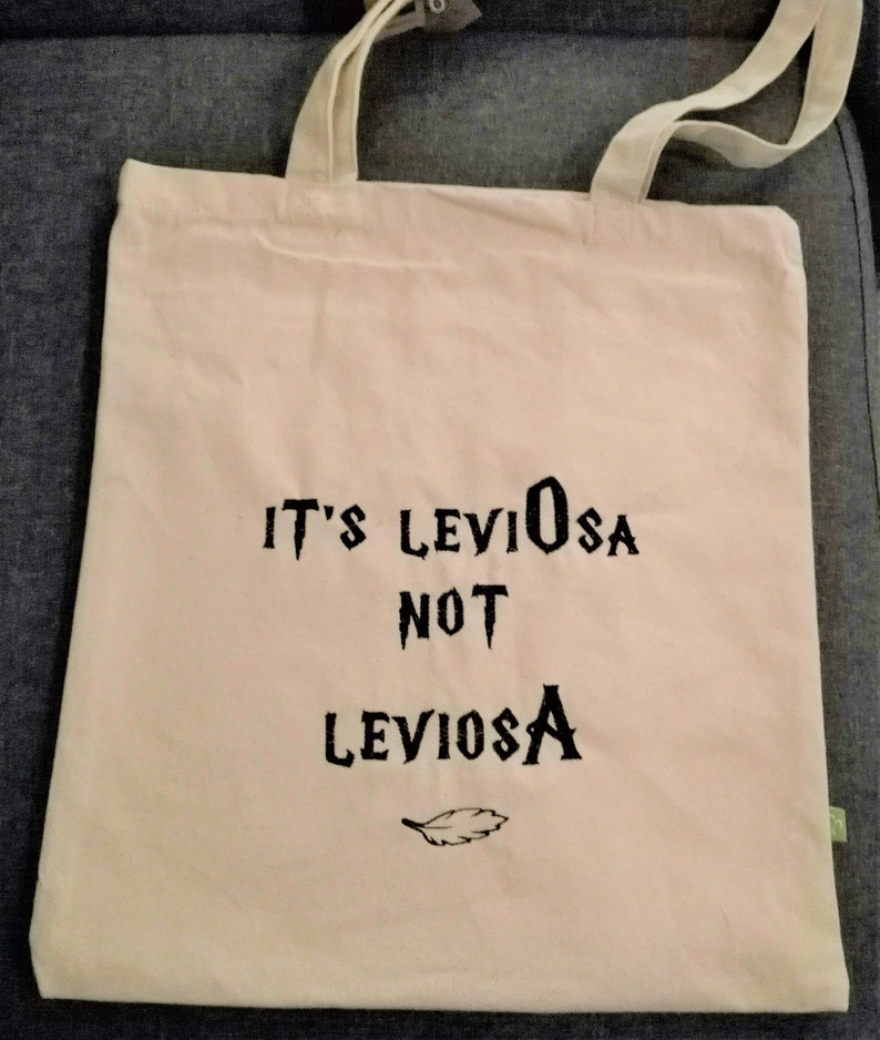 May include: A white canvas tote bag with black text that reads "It's LevioSA not LevioSA" and a small feather graphic.