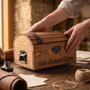 May include: A wooden chest with a curved lid, featuring dark metal latches, leather straps with studs, and the engraved name Luke Richard Ekstrom on the front. The box has a natural wood grain finish and is constructed with horizontal slats.