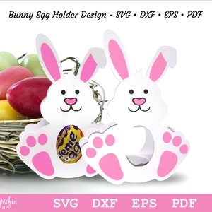 May include: Two white bunny shaped egg holders with pink ears and paws. The bunny's face has black eyes and a pink nose. The bunny's body has a cut-out to hold an egg. The bunny is sitting on a nest of straw.