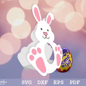 May include: White bunny shaped cardboard box with pink feet and a cut-out for an Easter egg. The box is sitting on a purple and white background. A purple and yellow chocolate egg is sitting in front of the box.