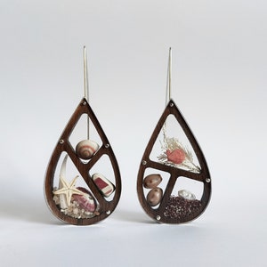 May include: A pair of teardrop earrings. Each earring has a dark wood frame with a clear centre, displaying miniature seashells, a starfish, and small stones. The earrings have silver-coloured ear wires.